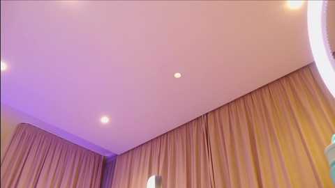 Media: Video of a modern room with beige curtains, light purple ceiling lights, and a circular mirror on the right. The room has a clean, minimalist design with soft lighting, creating a warm ambiance.