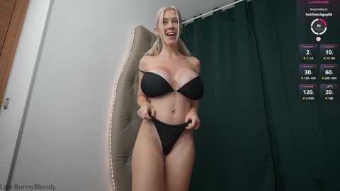 Media: Video of a smiling, curvy blonde woman with fair skin in black lingerie, standing in front of a beige upholstered chair and green curtain.