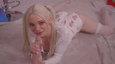 Media: Video of a blonde, fair-skinned woman with pigtails, wearing a white lace top and short white skirt, lying on her stomach on a bed, holding a dildo.