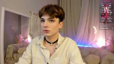 Media: Video of a young, slender, fair-skinned man with short, brown hair, wearing a white button-up shirt and black choker. Background features a cozy room with a bed, wall hanging, and dim lighting.