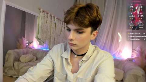 Media: Video of a young man with short brown hair, wearing a white button-down shirt and black choker, sitting in a dimly lit living room with a beige couch, hanging macrame, and a glowing fireplace in the background.