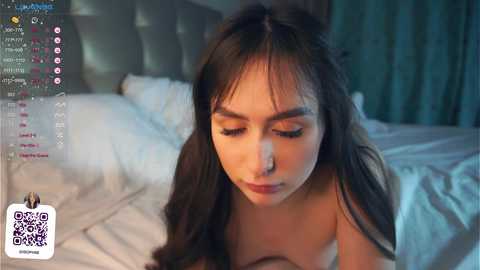 Media: A video of a young woman with long dark hair and closed eyes, lying on a bed with white sheets, in a dimly lit room.