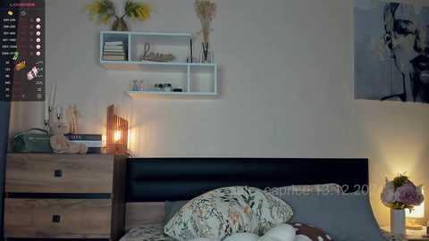 Media: Video of a cozy, minimalist bedroom with a wooden dresser, shelves, and a bed with floral bedding. A framed painting and a vase of flowers add warmth.