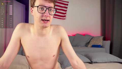 Media: Video of a young, slender, fair-skinned, shirtless man with short, light brown hair, wearing glasses, seated on a bed with gray sheets and pillows. An American flag hangs on the wall behind him.