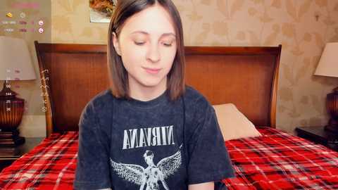 Media: Video of a young woman with straight brown hair, wearing a Nirvana t-shirt, sitting on a red and white plaid bedspread in a cozy bedroom with wooden headboard, floral wallpaper, and bedside lamps.