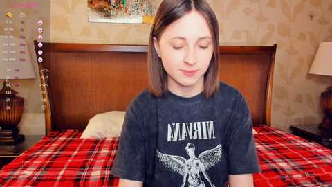 Video of a young, fair-skinned girl with shoulder-length brown hair wearing a Nirvana t-shirt, sitting on a red and black plaid bedspread in a cozy bedroom with vintage wallpaper and a wooden headboard.