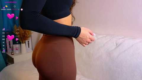 Media: A video of a woman in tight, high-waisted brown leggings and a black crop top, standing in a minimalist room with a white couch and a teal wall.