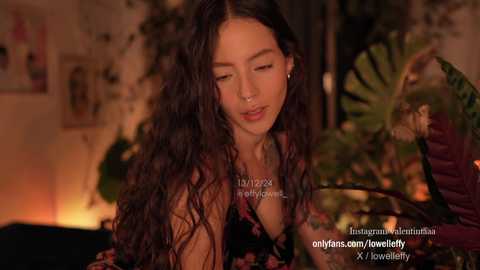 Media: A video of a young woman with long, curly black hair, wearing a black dress, sitting indoors near a fireplace, surrounded by green plants, in a dimly lit room.