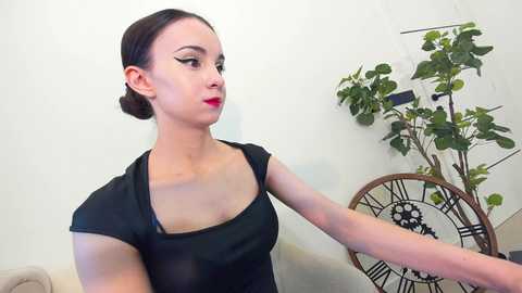 Media: Video of a young woman with fair skin, dark hair in a bun, wearing black clothing and red lipstick, in an indoor setting with a white wall and green plant.