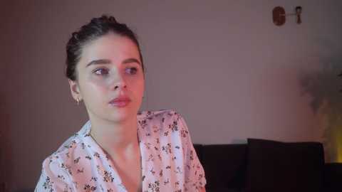 Media: Video of a young woman with dark hair in a bun, wearing a white floral robe, sitting on a dark couch against a plain wall, looking contemplative.