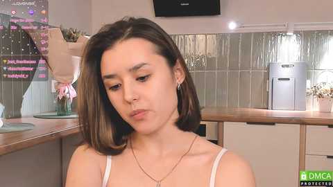 Media: Video of a young woman with straight, shoulder-length brown hair, wearing a white tank top, sitting in a modern kitchen with tiled walls, countertop, and a glass vase with flowers.