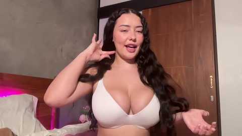 Media: A video of a curvy, light-skinned woman with long, wavy black hair, wearing a white bra, smiling and making a peace sign, standing in a modern bedroom with wooden furniture and a pink light.