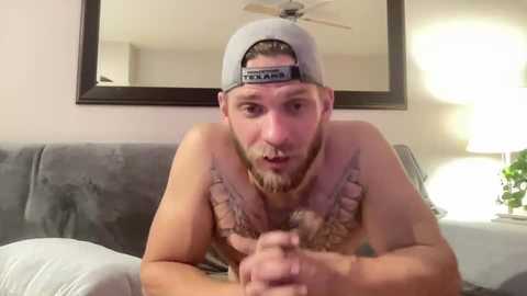 Media: Video of a shirtless man with a grey cap and tattoos on his chest, holding a beer bottle, sitting on a grey couch, in a dimly lit room with a table lamp.