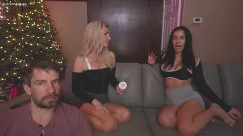 Media: Video of three people on a couch: a blonde woman in a black top, a brunette in a crop top and high-waisted shorts, and a bearded man in a brown shirt. Christmas tree and warm lighting in the background.