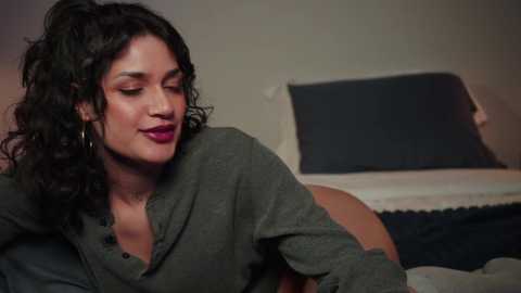 Media: Video of a smiling woman with curly dark hair, wearing a green shirt, sitting on a bed with a blue pillow and white blanket.