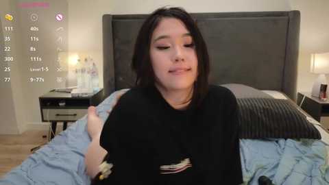 Media: Video of a young woman with shoulder-length black hair, wearing a black shirt, sitting on a bed with a gray headboard. She gives a thumbs-up gesture. The room has a modern, minimalist decor with beige walls and a nightstand with a lamp.