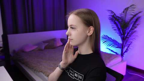 Media: Video of a young Caucasian woman with straight, light brown hair, wearing a black t-shirt, standing in a modern bedroom with purple lighting and a potted palm plant.