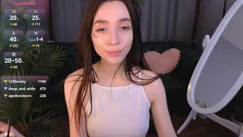 Media: A video of a young woman with long dark hair, fair skin, and a slender build, wearing a white tank top. She's indoors, near a mirror with a heart-shaped pillow in the background.