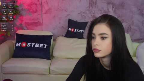 Media: Video of a young woman with long black hair and fair skin, wearing a black top, sitting on a beige sofa. The background features a dark blue cushion with \"MSTBET\" text and a purple wall.