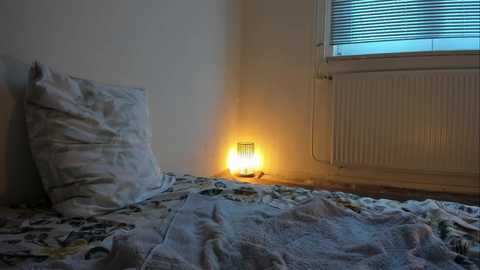 Media: Video of a dimly lit bedroom corner featuring a bed with white pillow and comforter, a small lit candle on the floor, and a radiator against the wall.