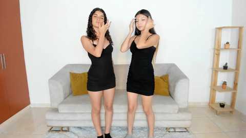 Media: Video of two young women with long dark hair, wearing tight black dresses, standing in front of a beige couch with mustard pillows, in a minimalist living room with light walls and wooden shelves.