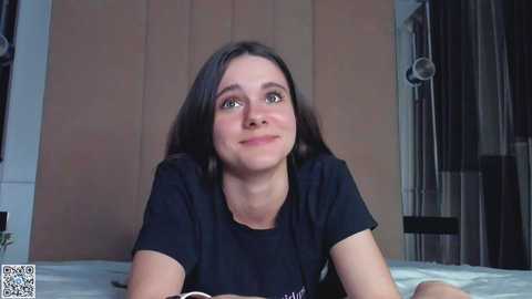 Media: Video of a young woman with long, dark hair, wearing a black t-shirt, lying on a bed in a dimly lit room with beige curtains and wooden wardrobe.