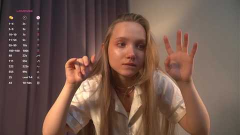 Media: Video of a young Caucasian woman with long blonde hair, wearing a white button-up shirt, making peace signs in front of a dark curtain background.