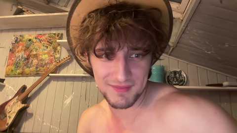 Media: Video of a shirtless young man with messy brown hair, wearing a beige cowboy hat, in a rustic room with a colorful painting, guitar, and a turquoise vase on a white shelf.