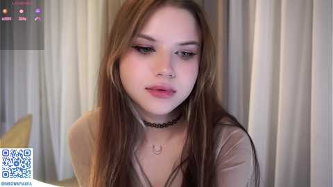 Video of a young, pale-skinned woman with long brown hair and subtle makeup, wearing a black choker and beige top, sitting against a beige curtain background.