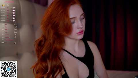 Media: Video of a fair-skinned woman with long, wavy red hair, wearing a black tank top, sitting in a dimly lit room with dark curtains.