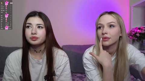 Media: Video of two young women with long hair, one brunette and one blonde, seated on a couch, both wearing white tops. Background includes a purple light, flowers, and a starry wall decoration.