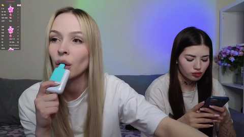 Media: Video of two young women with long hair, one blonde and one brunette, both holding blue and white Nintendo Game Boys, sitting on a couch in a dimly lit room.