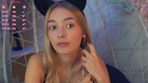 Media: Video of a young blonde woman with fair skin, wearing a black headband and necklace, indoors with a blurred background.