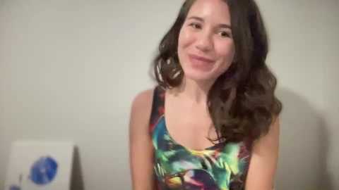 Media: Video of a young woman with long, wavy brown hair, fair skin, and a slim figure, wearing a colorful, patterned tank top, smiling against a plain, light-colored wall.