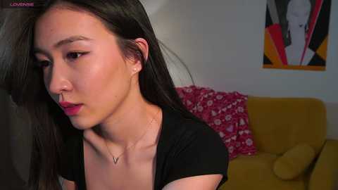 Media: A video of a young Asian woman with long black hair, wearing a black top and nose ring, sitting on a mustard-yellow couch with red floral pillows in a modern, minimalist living room.