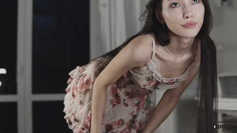 Media: Video of a slender, young Asian woman with long black hair, wearing a floral dress, leaning forward, indoors at night, window visible in the background.