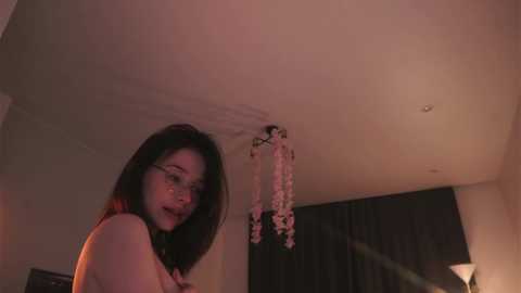 Media: Video of an Asian woman with long black hair, glasses, and pink lips, wearing a black top, standing in a dimly lit room with a hanging flower arrangement and a lamp.