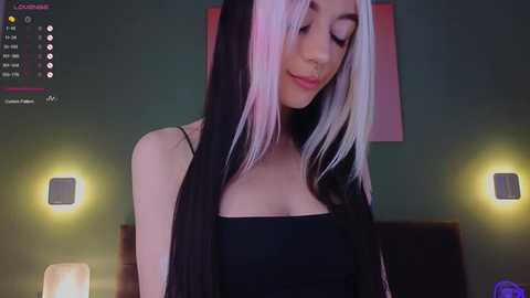 Media: Video of a young woman with long black and white hair, wearing a black spaghetti-strap top, standing in a dimly lit room with green walls and two modern lamps.