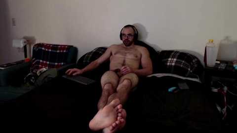 Media: Video of a nude man with a beard, wearing headphones, lying on a bed with black sheets and plaid pillows, surrounded by clutter.