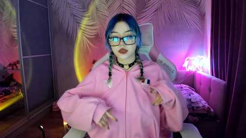 Media: A young woman with blue hair and glasses, wearing a pink hoodie, poses in a brightly lit, tropical-themed bedroom.