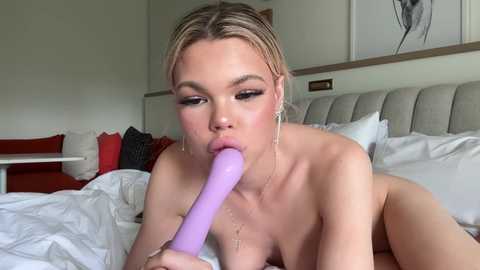 Media: Video of a nude blonde woman with fair skin, medium breasts, and a slender physique, performing oral sex with a lavender vibrator on a bed with white sheets and pillows, in a modern, minimalist bedroom with white walls and a large headboard.