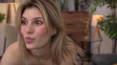 Media: Video of a fair-skinned woman with shoulder-length blonde hair, wearing a gold necklace, making a kissing face in a softly lit room with green plants and wooden furniture.