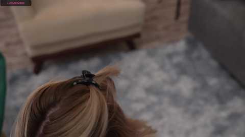 Media: A video of a blonde woman with a black scrunchie in her hair, sitting on a patterned rug in a cozy living room with beige furniture and a brick fireplace in the background.