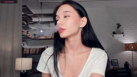 Video of an East Asian woman with long black hair, wearing a white V-neck top, exhaling cigarette smoke in a dimly lit, modern living room with bookshelves and a lamp.