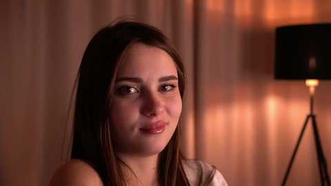 Media: A video of a young woman with straight, dark brown hair, fair skin, and a soft smile, seated in a warmly lit room with beige curtains and a black lamp.