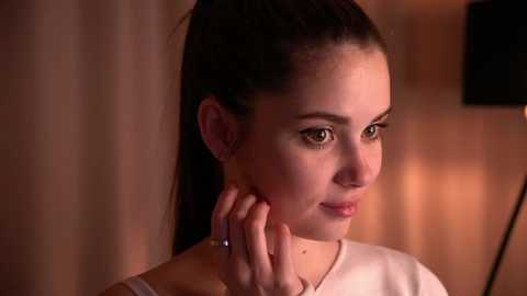 Media: Video of a young woman with fair skin, dark hair pulled back, wearing a white top, standing indoors with warm lighting. She gazes thoughtfully, hand touching her cheek.