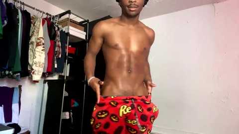 Media: Video of a shirtless, muscular Black man with dark skin, wearing red Batman-printed pajama pants, standing in a cluttered, dimly lit room with hanging clothes and a white wall.