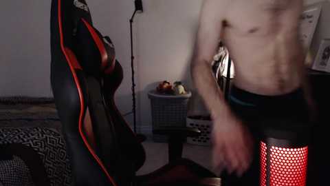 Media: Video of a shirtless, lean man in black shorts standing next to a red and black gaming chair with a basket of fruits in a dimly lit room.