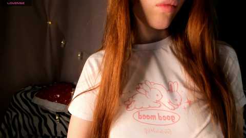 Video of a fair-skinned woman with long red hair, wearing a white T-shirt with a \"boom boob\" design, in a dimly lit bedroom with zebra-print bedding.