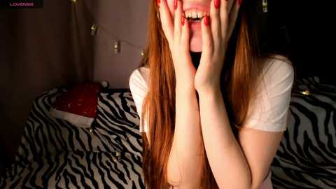 Video of a young woman with long, straight red hair, fair skin, and a white T-shirt, covering her face with both hands, sitting on a zebra-patterned bed.